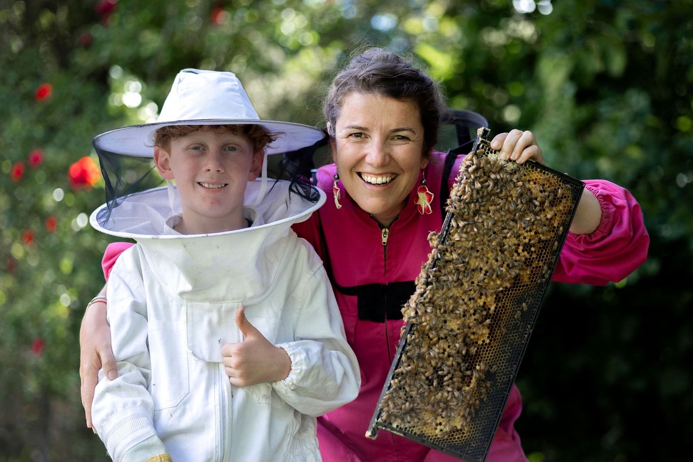 A beekeeper and a young student in a bee suit proudly holding a honeycomb frame covered in bees